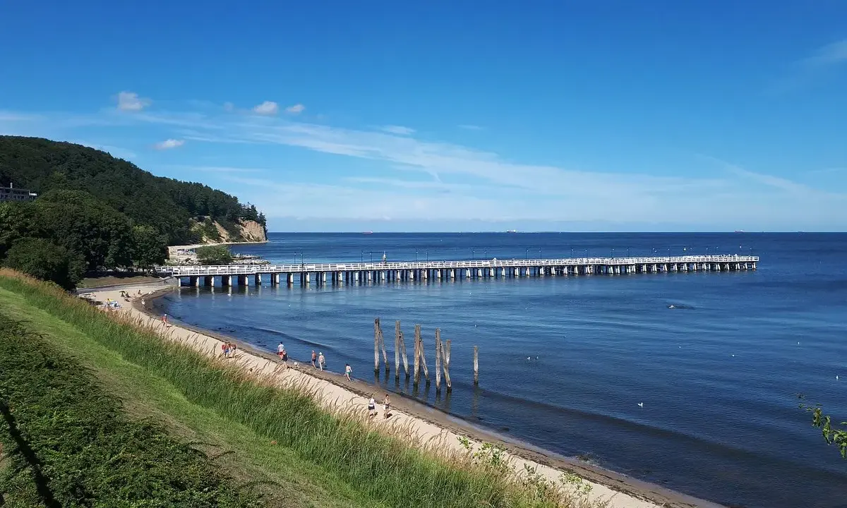 En lång pir sträcker sig ut i det blå havet vid Gdynia, Polen. En grön kulle med träd reser sig vid sidan av stranden där några människor promenerar.