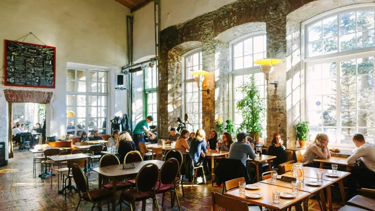 Folk äter och dricker på en livlig restaurang i Tallinn. Stora fönster släpper in solljus i lokalen med rustika tegelväggar.