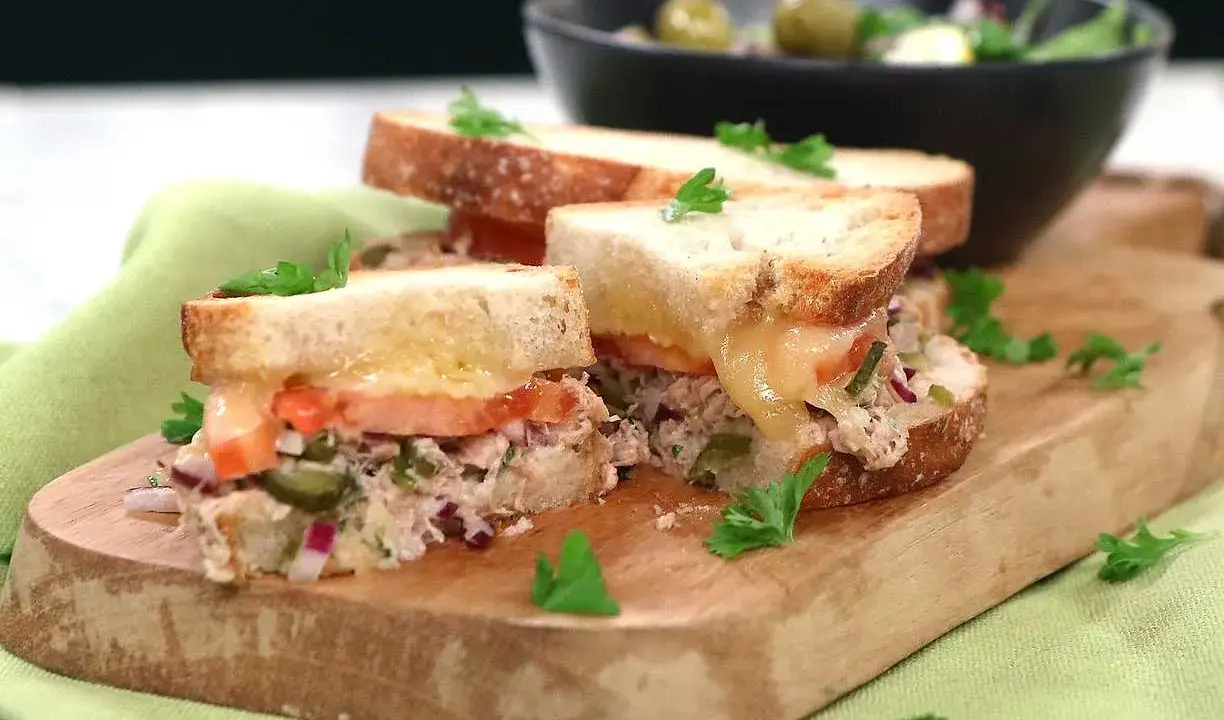 Två halvor av en gyllene tuna melt med smält ost, tomat och kapris, garnerad med persilja. Serveras på en träskärbräda med en sallad i bakgrunden.