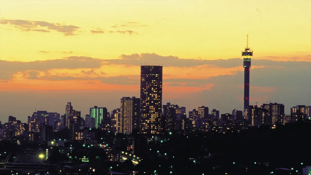 Stadssilhuett i Johannesburg, Sydafrika, med upplysta skyskrapor och ett högt torn mot en gul och orange solnedgångshimmel.