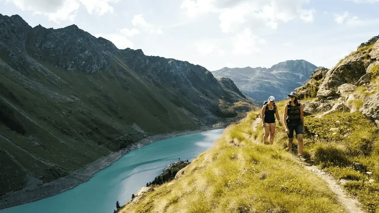 Två personer vandrar på en bergstig med en turkos sjö och dramatiska berg i bakgrunden. En perfekt dag för att vandra i Schweiz.
