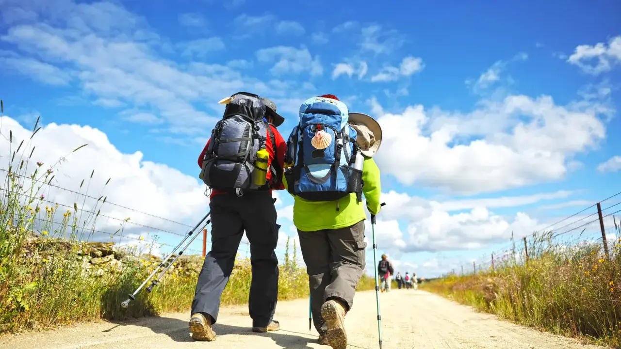 Två vandrare med ryggsäckar och vandringsstavar går längs en grusväg under en blå himmel med moln. Kanske en del av pilgrimsleder Sverige.
