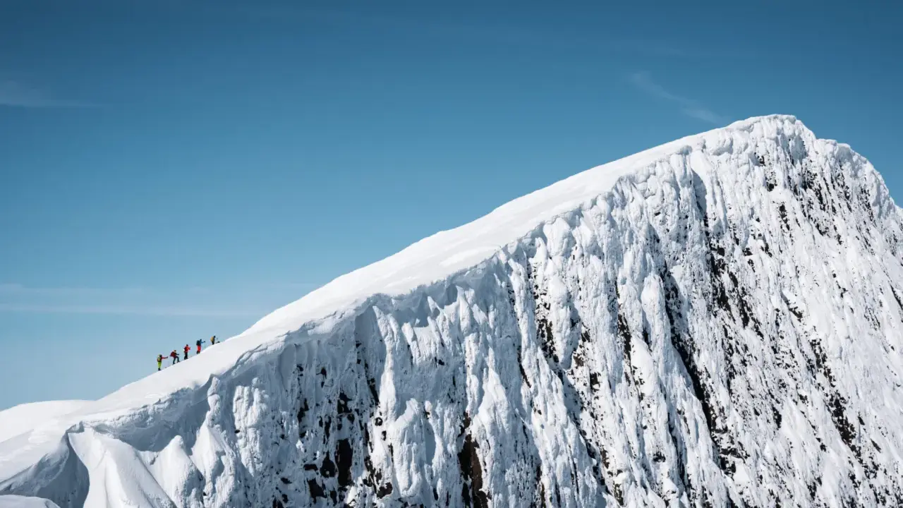 En grupp vandrare på en snöig bergstopp under en helags topptur. Den isiga bergväggen reser sig dramatiskt mot en klarblå himmel.