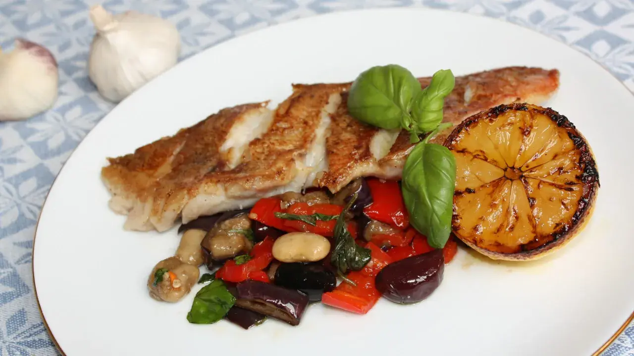 Pan-seared fish with roasted vegetables, a grilled lemon slice, and basil on a white plate.