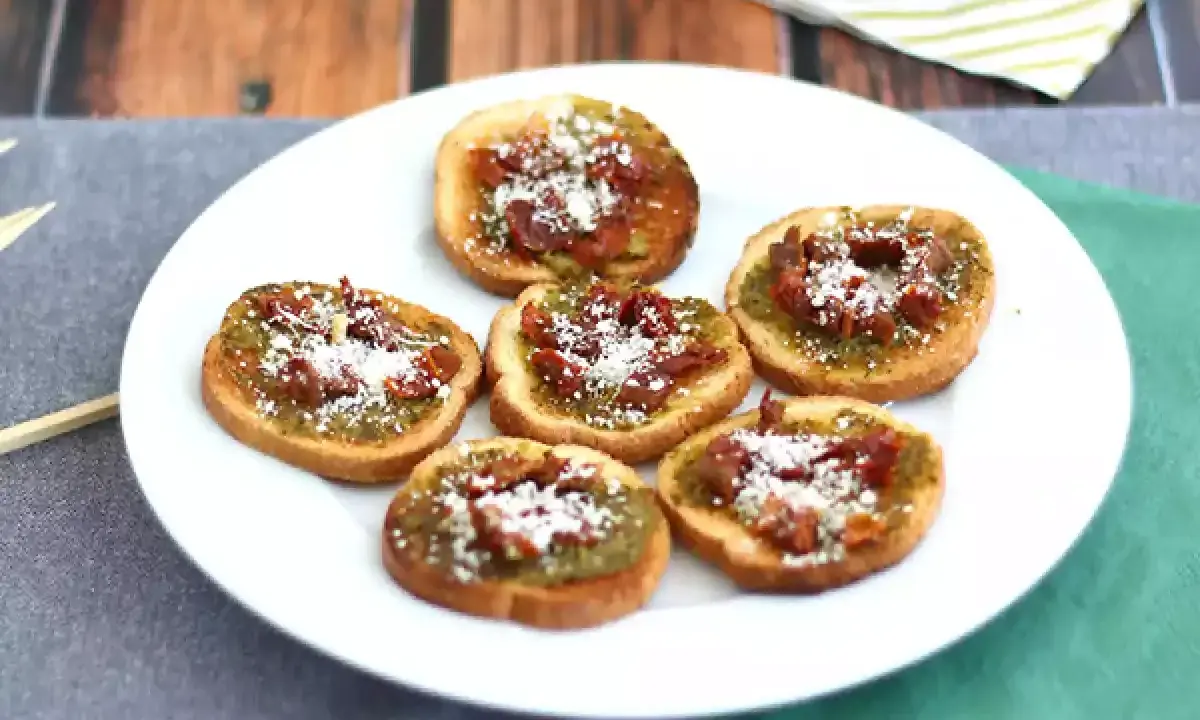 Små, rostade brödskivor toppade med grönt pesto, soltorkade tomater och riven ost. Perfekt som en enkel pesto toast.