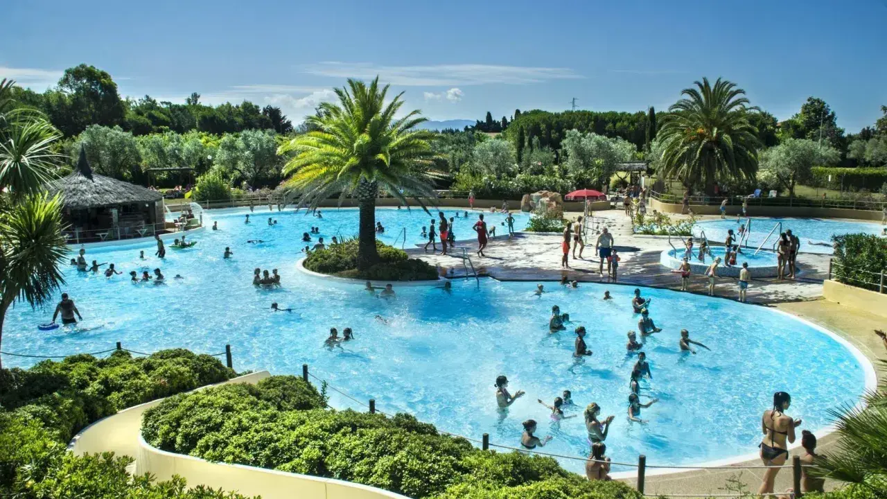 En stor pool med många badande människor, palmer och en vattenrutschkana. En härlig semesterbild från ett camping i Italien.