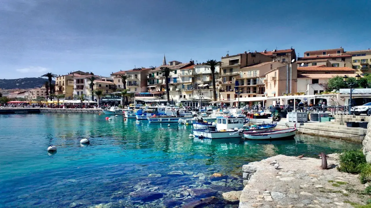 Båtar i en turkos hamn i Calvi, Korsika, med färgglada hus och palmer längs kajen.