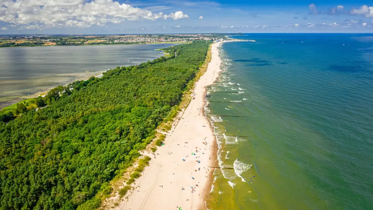 En lång sandstrand kantad av en grönskande skog. Havet glittrar i solljuset och en stad syns i bakgrunden. En perfekt sommardag på hel polen.