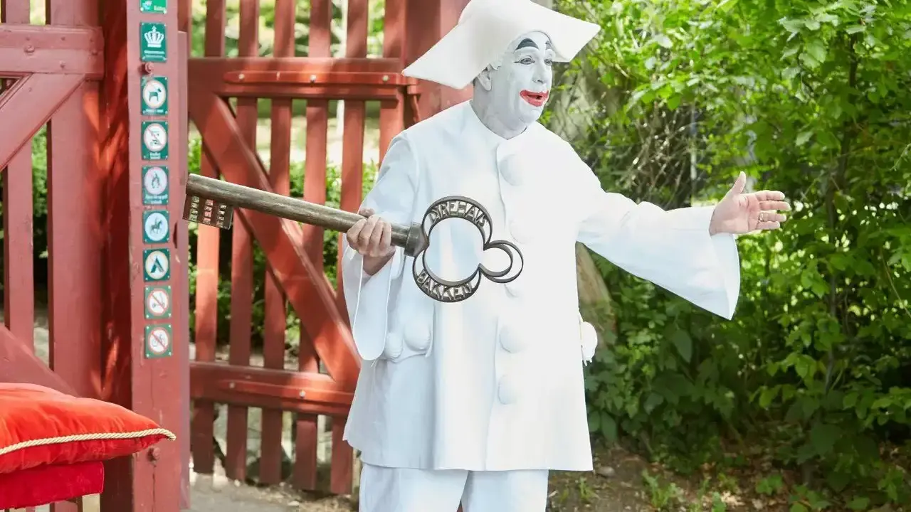 En clown med vit smink och stor hatt håller en nyckel med texten "Dyrehavs Bakken". Han står vid en röd grind.