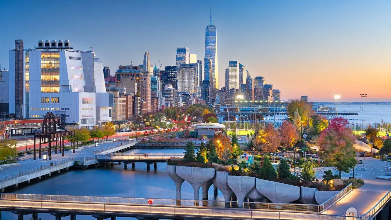 Fakta om New York: En vy över Manhattans skyline vid solnedgången, med den ikoniska One World Trade Center och en modern park vid vattnet.