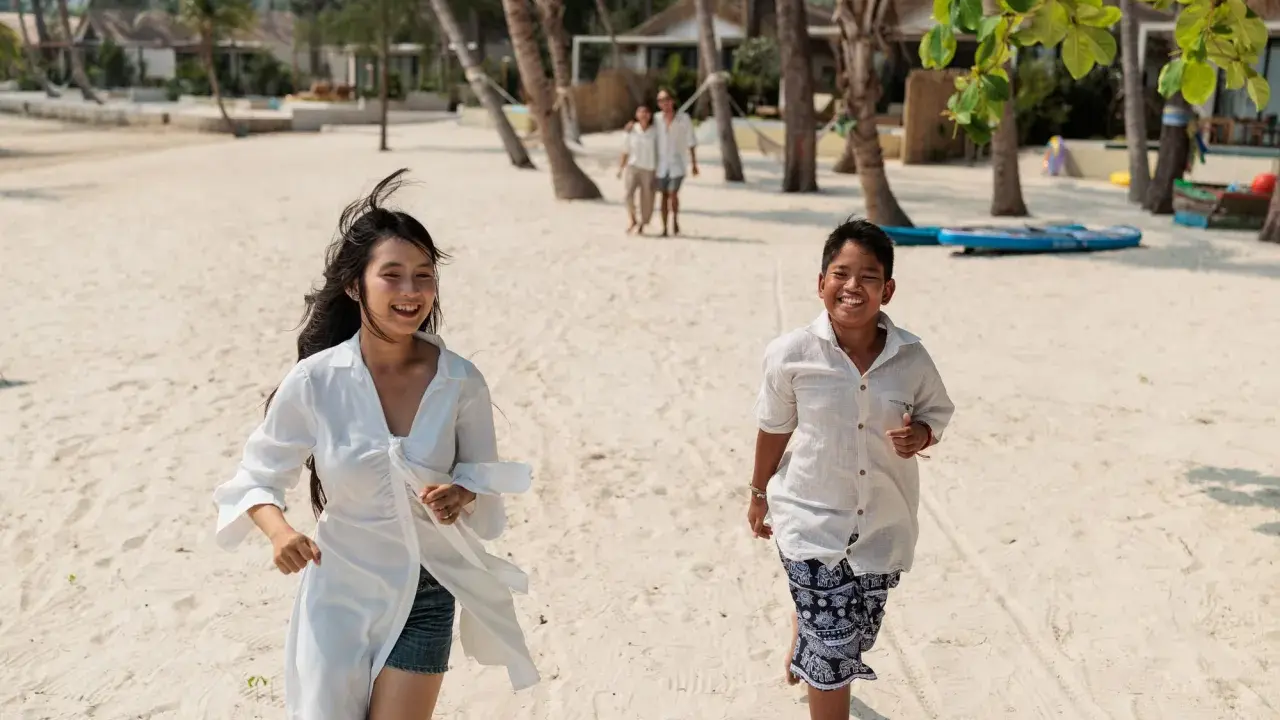 Två barn springer glatt på en sandstrand i Thailand. I bakgrunden syns palmer och bungalows.