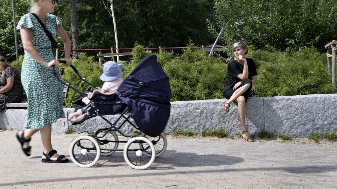 En kvinna med barnvagn njuter av sin semester i Sverige med barn. En annan kvinna sitter på en stenmur i bakgrunden.