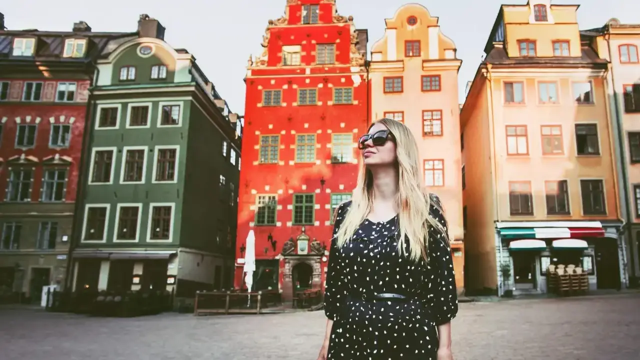 En kvinna med solglasögon står framför färgglada hus på ett torg. Kanske är detta sveriges vackraste stad?