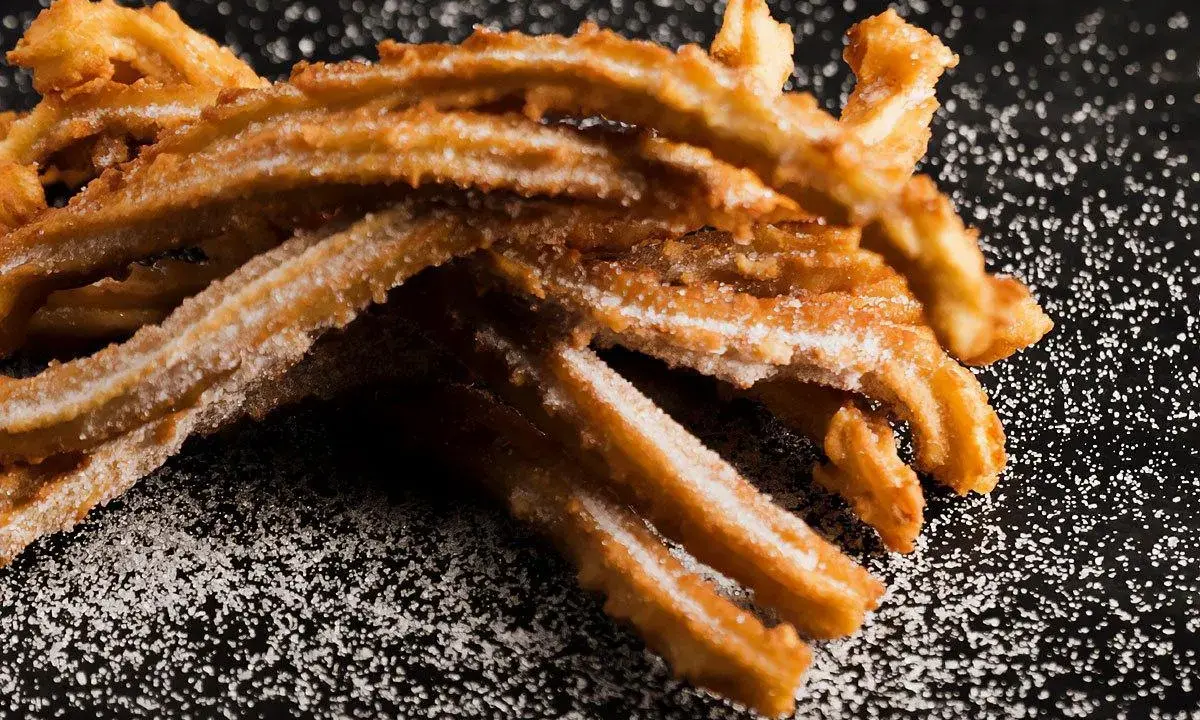 En hög med gyllene, sockerbeströdda churros, en härlig friterad deg, ligger på en mörk yta.