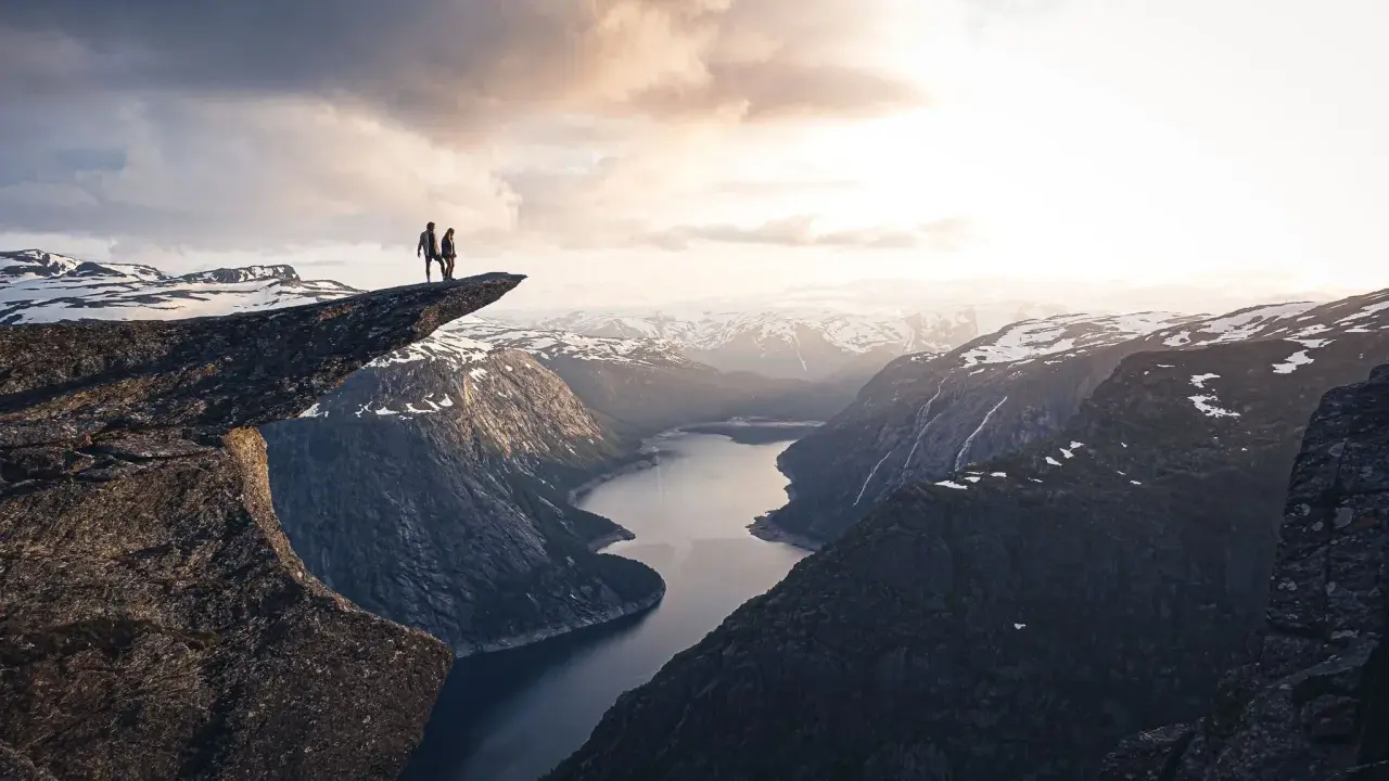 Två personer står på Trolltunga efter en 3 dagars vandring i Norge. Dramatisk utsikt över fjord och snöklädda berg.