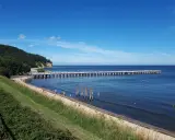 En lång pir sträcker sig ut i det blå havet vid Gdynia, Polen. En grön kulle med träd reser sig vid sidan av stranden där några människor promenerar.