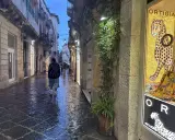 En person går längs en regnvåt gata i Syrakusa, Sicilien. En butiks skyltfönster visar en mosaik av en jaguar och texten "Ortigia".