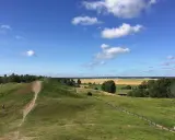 Gröna kullar med stigar och en ensam person. Ett vackert landskap att se i Uppsala med fält och skog i bakgrunden under en blå himmel med moln.