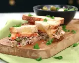Två halvor av en gyllene tuna melt med smält ost, tomat och kapris, garnerad med persilja. Serveras på en träskärbräda med en sallad i bakgrunden.