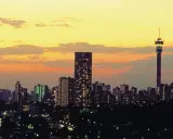 Stadssilhuett i Johannesburg, Sydafrika, med upplysta skyskrapor och ett högt torn mot en gul och orange solnedgångshimmel.