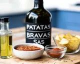 Ingredienser för patatas bravas recept: en flaska patatas bravas sås, olivolja, två skålar med sås och en skål med potatis.