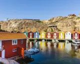 Färgglada sjöbodar längs en klippig kust, perfekt för en weekendresa Sverige. Båtar guppar i det spegelblanka vattnet.