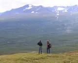 Två vandrare njuter av utsikten över vidsträckta fjäll och skogar, kanske under en vandra abisko 3 dagar. Snöklädda berg tornar upp sig i bakgrunden.