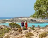 Ett par njuter av utsikten över Elafonissi, Kreta, med dess turkosa vatten och steniga landskap. En röd fallskärm syns i fjärran.