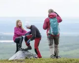 Två barn hjälper varandra upp på en sten under en vandring. En vuxen står bakom med ryggsäcken redo för äventyr.