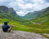 Person sitter på en sten och njuter av utsikten över en grön dal i Skottland. En perfekt plats för att vandra i Skottland.