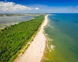 En lång sandstrand kantad av en grönskande skog. Havet glittrar i solljuset och en stad syns i bakgrunden. En perfekt sommardag på hel polen.