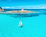 En segelbåt glider fram på turkost vatten mot en gammal vakttorn på en ö. Perfekt för din resa till Sardinien tips!