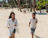 Två barn springer glatt på en sandstrand i Thailand. I bakgrunden syns palmer och bungalows.