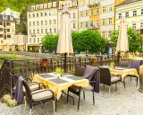 Uteservering vid en flod i Karlovy Vary med gula byggnader i bakgrunden.