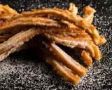 En hög med gyllene, sockerbeströdda churros, en härlig friterad deg, ligger på en mörk yta.
