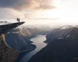 Två personer står på Trolltunga efter en 3 dagars vandring i Norge. Dramatisk utsikt över fjord och snöklädda berg.