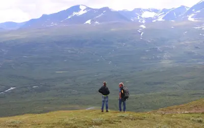 Två vandrare njuter av utsikten över vidsträckta fjäll och skogar, kanske under en vandra abisko 3 dagar. Snöklädda berg tornar upp sig i bakgrunden.