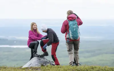 Två barn hjälper varandra upp på en sten under en vandring. En vuxen står bakom med ryggsäcken redo för äventyr.