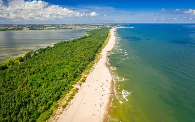 En lång sandstrand kantad av en grönskande skog. Havet glittrar i solljuset och en stad syns i bakgrunden. En perfekt sommardag på hel polen.