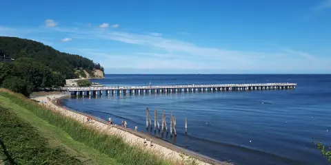En lång pir sträcker sig ut i det blå havet vid Gdynia, Polen. En grön kulle med träd reser sig vid sidan av stranden där några människor promenerar.
