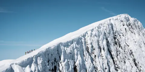 En grupp vandrare på en snöig bergstopp under en helags topptur. Den isiga bergväggen reser sig dramatiskt mot en klarblå himmel.