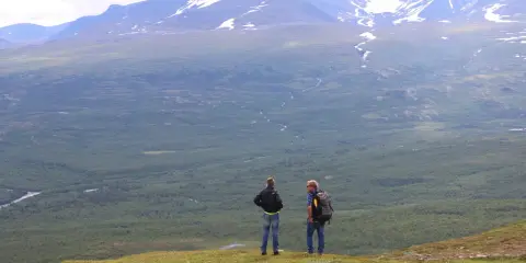 Två vandrare njuter av utsikten över vidsträckta fjäll och skogar, kanske under en vandra abisko 3 dagar. Snöklädda berg tornar upp sig i bakgrunden.