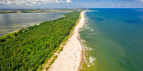 En lång sandstrand kantad av en grönskande skog. Havet glittrar i solljuset och en stad syns i bakgrunden. En perfekt sommardag på hel polen.