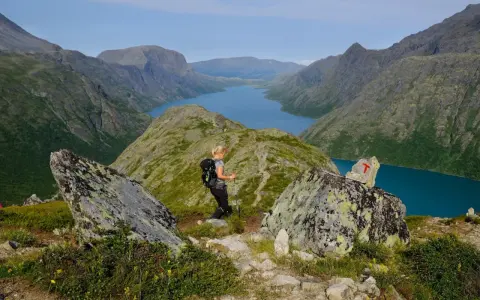 En kvinna vandrar i Jotunheimen med en blå sjö och berg i bakgrunden. En T-märkt sten visar vägen.