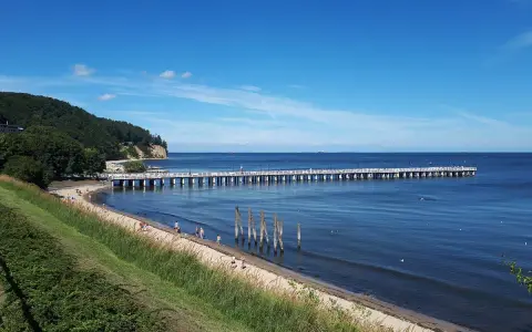 En lång pir sträcker sig ut i det blå havet vid Gdynia, Polen. En grön kulle med träd reser sig vid sidan av stranden där några människor promenerar.