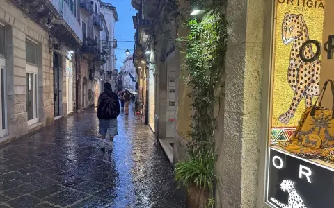 En person går längs en regnvåt gata i Syrakusa, Sicilien. En butiks skyltfönster visar en mosaik av en jaguar och texten "Ortigia".