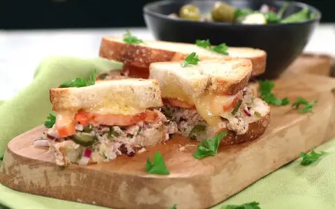 Två halvor av en gyllene tuna melt med smält ost, tomat och kapris, garnerad med persilja. Serveras på en träskärbräda med en sallad i bakgrunden.