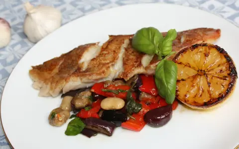 Pan-seared fish with roasted vegetables, a grilled lemon slice, and basil on a white plate.