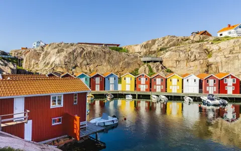 Färgglada sjöbodar längs en klippig kust, perfekt för en weekendresa Sverige. Båtar guppar i det spegelblanka vattnet.