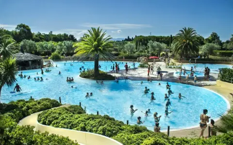 En stor pool med många badande människor, palmer och en vattenrutschkana. En härlig semesterbild från ett camping i Italien.